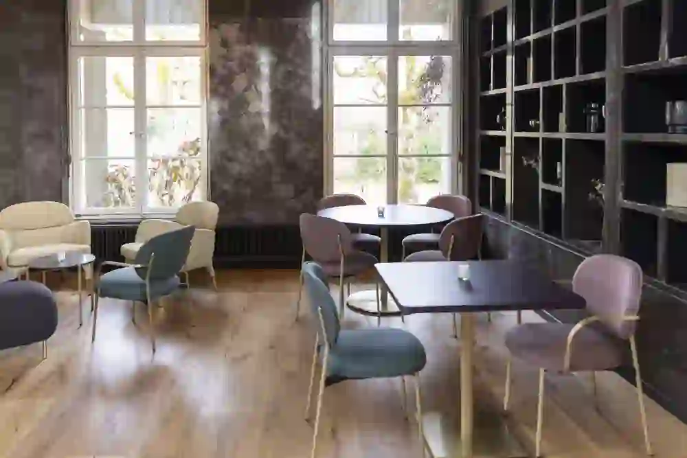 accogliente salotto con poltrone imbottite nei colori beige, rosa e petrolio, tavolini rotondi e pavimento in legno. Le grandi finestre lasciano entrare molta luce, mentre una parete scura di scaffali crea un elegante contrasto.