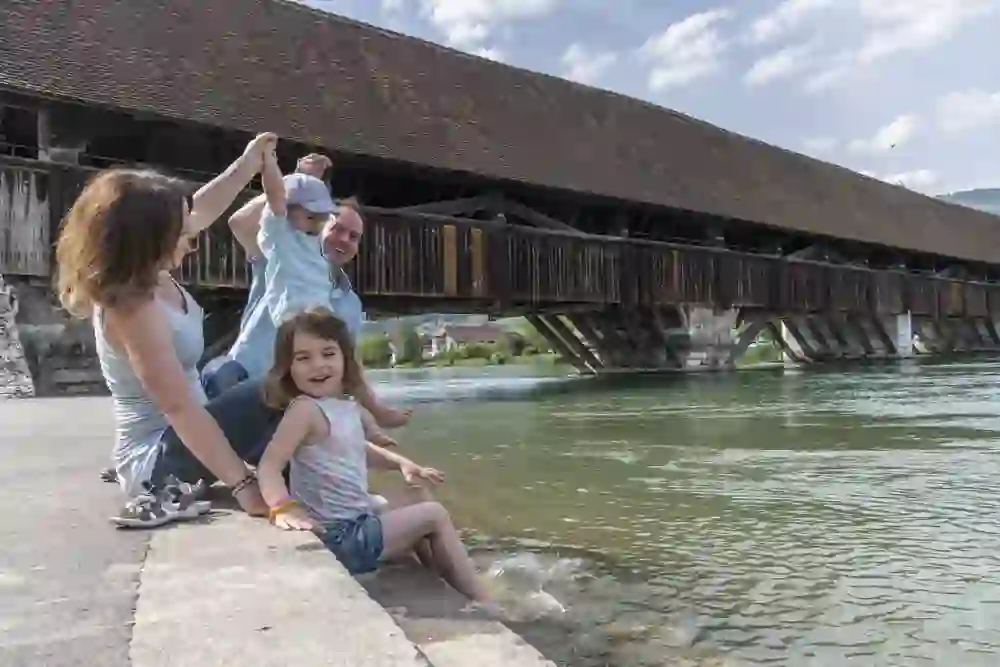 Eine Familie geniesst eine sorglose Zeit am Ufer bei Wangen an der Aare; die Kinder spielen im Wasser, während die Eltern liebevoll zuschauen, umgeben von der malerischen Kulisse der alten Holzbrücke und der ruhigen Aare.
