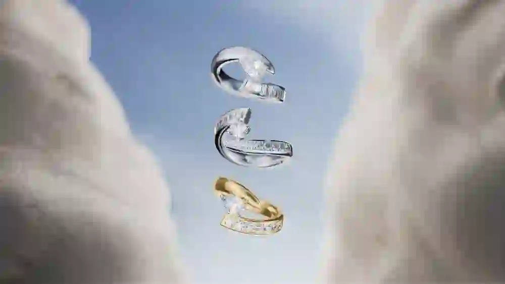 Die Aufnahme zeigt drei luxuriöse Ringe von Bucherer, die scheinbar schwerelos zwischen zwei weich verschwommenen, stoffartigen Formen in einem wolkenartigen Umfeld schweben. Die Ringe – zwei aus Weissgold, einer aus Gelbgold – sind mit funkelnden Brillanten und Baguette-Diamanten besetzt. Die luftige, lichtdurchflutete Inszenierung vor einem klaren Himmelshintergrund unterstreicht die Eleganz und Leichtigkeit des Designs und rückt die edlen Schmuckstücke in den Fokus.