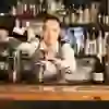 A smiling bartender pours concentrated liqueur into a measuring cup, surrounded by a lively bar with a wide selection of drinks.