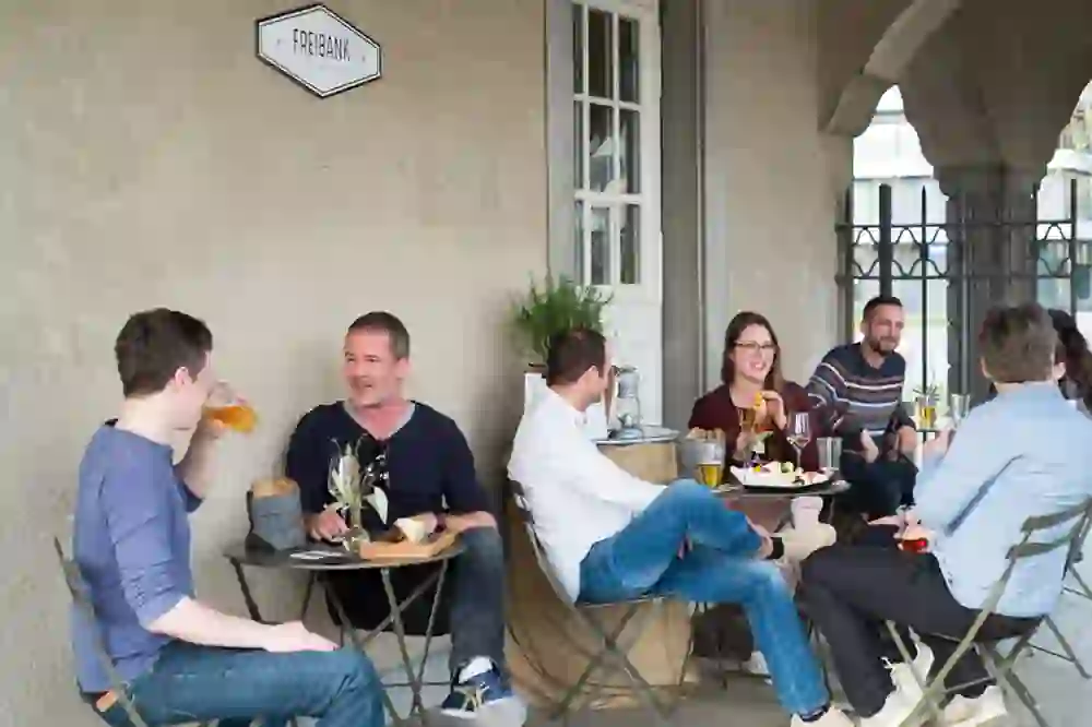 The picture shows a group of people sitting outside in a café or bar, apparently in a good mood. Some are enjoying drinks and it seems as if they are having animated conversations. The atmosphere is relaxed and sociable.