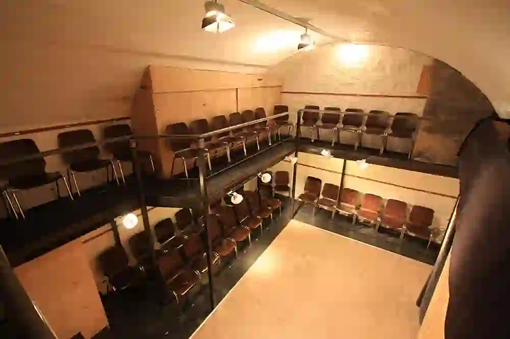 Das Foto zeigt das Zytglogge Theater mit einer intimen Tribüne. Die Sitzreihen sind in einem schlichten, funktionalen Design gehalten, das den historischen Charakter des Gewölberaums ergänzt. Die Beleuchtung ist einfach und fokussiert, was die Aufmerksamkeit auf die Bühnenfläche lenkt, die im Moment leer zu sein scheint und auf die nächste Aufführung wartet.