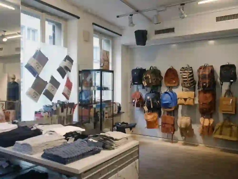 Auf dem Bild ist das Innere eines modernen Bekleidungsgeschäfts zu sehen. An den Wänden hängen verschiedene Rucksäcke, und es gibt ein Display mit gefalteten Hemden und Socken. Im Hintergrund steht eine Glasvitrine neben einem Schaufenster, durch das Tageslicht einfällt.