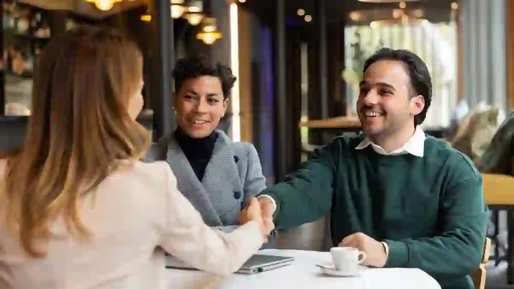 Herzlicher Business-Moment im stilvollen Café: Ein Mann begrüßt eine Frau per Handschlag, begleitet von einer Kollegin. Warme Lichtstimmung, entspannte Professionalität und vertrauensvolle Atmosphäre für Networking und Meetings.
