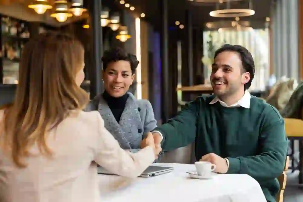 Un caloroso momento di business in un elegante caffè: un uomo saluta una donna con una stretta di mano, accompagnato da una collega donna. Luci calde, professionalità rilassata e un'atmosfera di fiducia per il networking e gli incontri.
