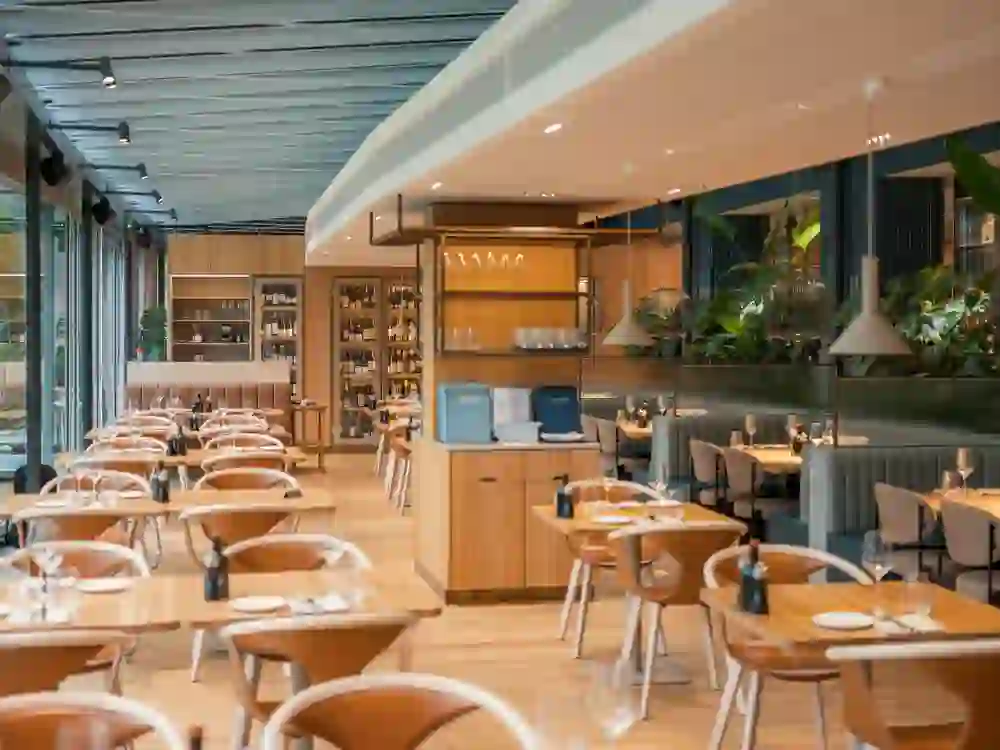 Das Bild zeigt ein elegantes Restaurant mit modernem Interieur, Holztischen und bequemen Sitzgelegenheiten. Natürliches Licht und Pflanzen schaffen eine frische, einladende Atmosphäre.