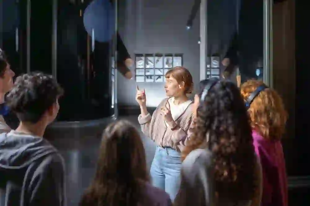 Une médiatrice explique l'exposition Moneyverse à un petit groupe. Les visiteurs écoutent attentivement tandis qu'elle montre une installation. La scène semble curieuse et vivante, marquée par l'envie de découvrir et d'aborder ensemble le thème de l'argent.