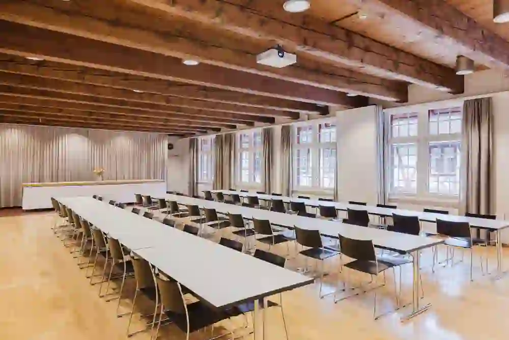 La salle donne une impression de calme, de clarté et de concentration. Les longues rangées de tables en forme de U créent une atmosphère structurée et propice au dialogue - idéale pour les réunions, les ateliers ou les séminaires. Les poutres en bois apparentes au plafond confèrent à la salle chaleur et caractère, tandis que les grandes fenêtres laissent entrer beaucoup de lumière du jour et rendent le contexte historique perceptible. Dans l'ensemble, il en résulte une interaction équilibrée entre fonctionnalité, discrétion et élégance intemporelle.