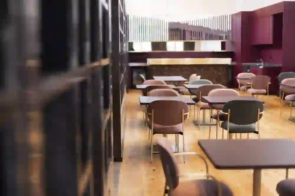 Vista della sala da pranzo con piccoli tavoli quadrati e sedie colorate. Sullo sfondo, il bar e una struttura a scaffale aperto, dall'eleganza moderna e dai colori caldi.