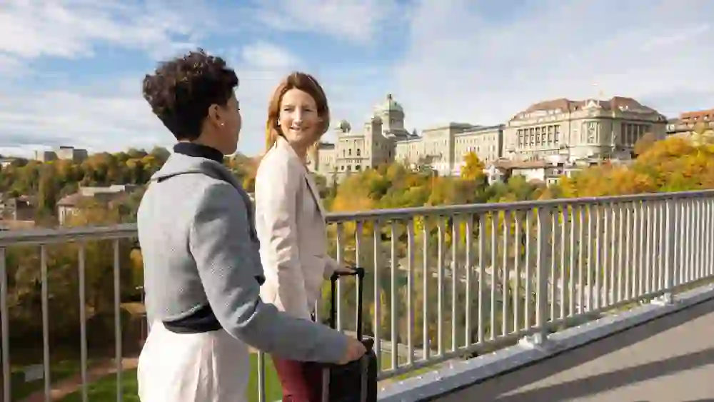 Zwei Reisende mit Rollkoffern auf der Kirchenfeldbrücke in Bern, mit Blick aufs Bundeshaus und herbstlich leuchtende Bäume. Die Stimmung ist entspannt, inspirierend und urban – perfekt für einen genussvollen Bleisure-Trip.