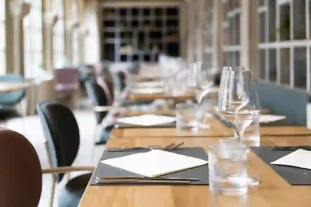 Luminoso giardino d'inverno con lunghi tavoli in legno e sedie imbottite lungo le grandi finestre. Gli alti archi e le mensole a muro strutturate conferiscono all'ambiente un'atmosfera elegante e architettonica.