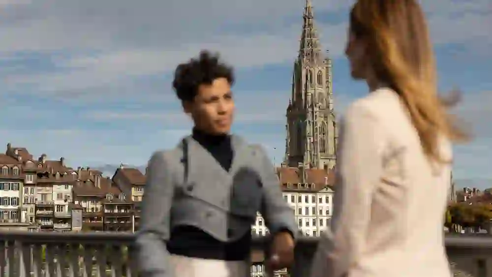 Zwei Frauen im Gespräch auf der Kirchenfeldbrücke in Bern, im Hintergrund Altstadt und Münster. Ruhige, stilvolle Bleisure-Atmosphäre mit urbanem Charme, sanftem Licht und Weitblick über die Dächer der Stadt.