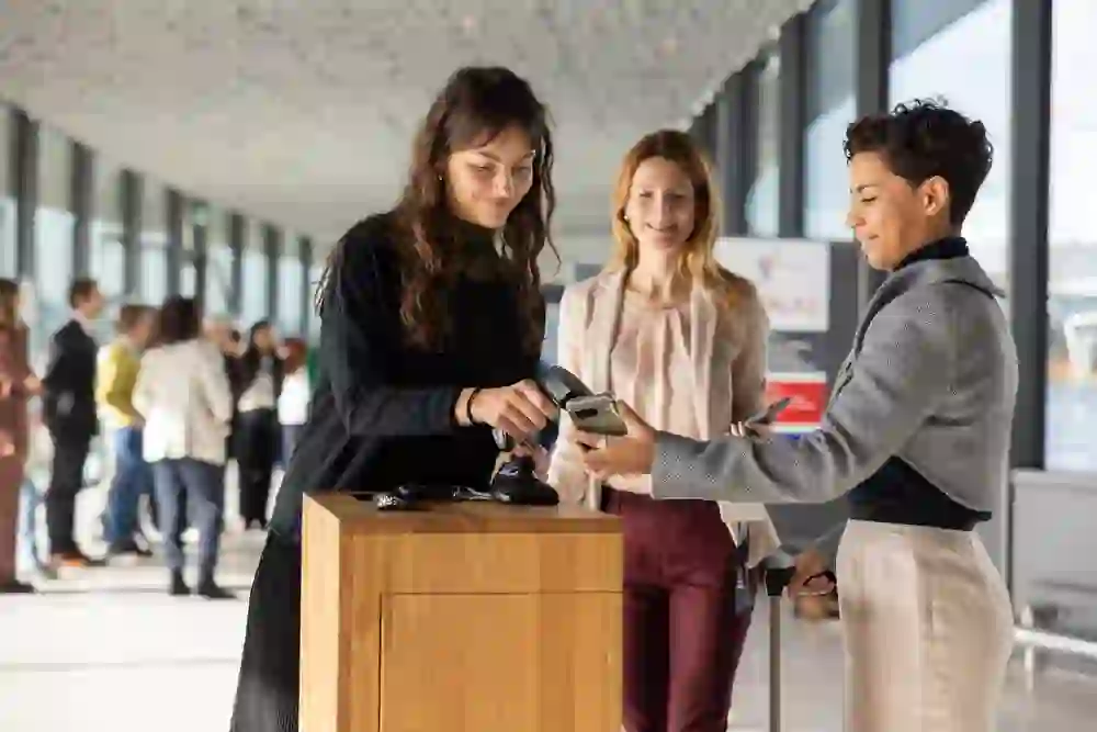 Freundlicher Empfang im lichtdurchfluteten Foyer: Eine Teilnehmerin checkt am Counter ein, während zwei Frauen lächelnd unterstützen. Moderne, professionelle Atmosphäre mit entspannter, einladender Stimmung und Networking-Flair.