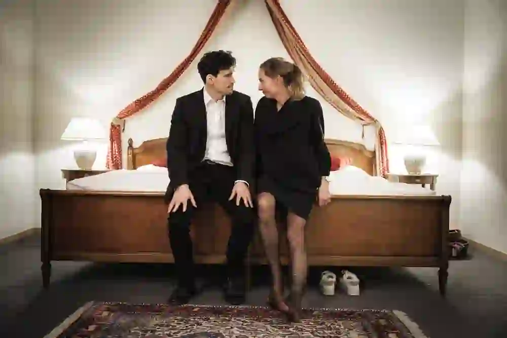 The picture shows a scene from the hotel room play "Toulouse" in the theatre on Effingerstrasse. An elegantly dressed couple is sitting on a bed and gazing intently at each other. The intimate, mysterious atmosphere between closeness and distance arouses curiosity and emotion.