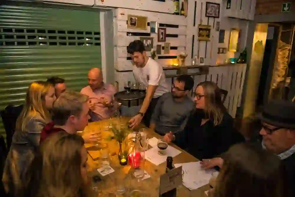 Das Foto fängt eine Gruppe von Menschen ein, die an einem gemütlichen Tisch in einem warm beleuchteten Raum mit einer rustikalen, dekorativen Wand sitzen. Man sieht leere Gläser und Flaschen, was darauf hindeutet, dass hier eine Bierverkostung stattfindet. Ein Mann im Vordergrund, vermutlich der Gastgeber oder Bierkenner, scheint etwas zu erklären oder zu diskutieren, während die anderen Teilnehmer interessiert zuhören oder sich unterhalten. Die Stimmung ist gesellig und entspannt.