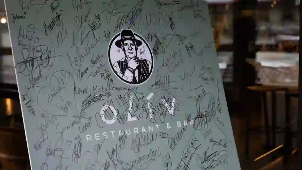 Sur l'image, on peut voir un panneau ou une affiche recouverte de nombreuses signatures et dédicaces. Au centre de la plaque se trouve un logo circulaire représentant un homme en costume et chapeau, avec la mention "OLIV Restaurant & Bar" en dessous. Les signatures varient en style et en taille, ce qui indique que de nombreuses personnes différentes ont signé le panneau. En arrière-plan, on distingue vaguement le restaurant.