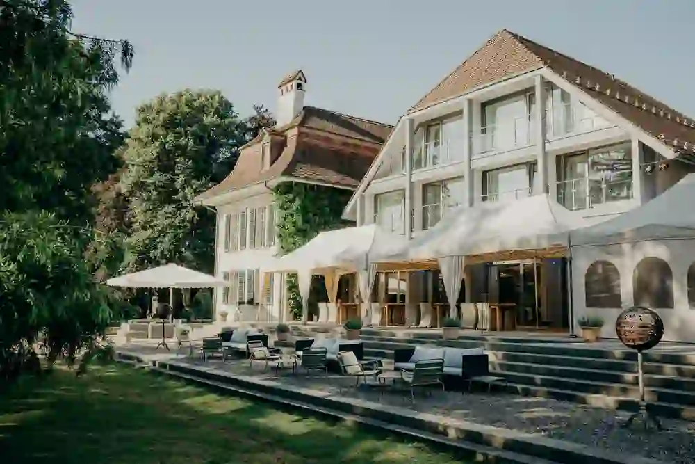 The picture shows an elegant outdoor terrace of a historic building. Parasols and cushioned seating invite you to linger outside. The scene is peaceful and inviting, with a manicured lawn and surrounded by rich greenery. The steps lead the eye to the modern extension of the traditional building, suggesting a harmonious blend of old and new. The atmosphere is calm, almost idyllic - perfect for a relaxing break or a social gathering.