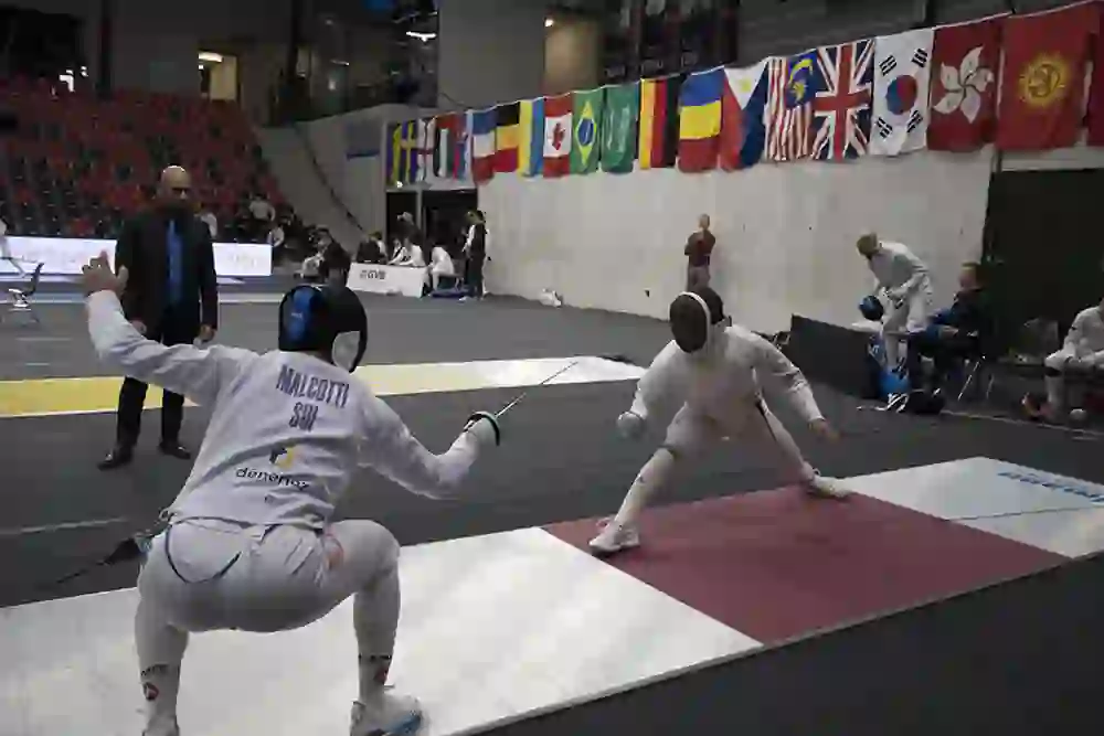 L'immagine mostra un emozionante duello di spada. Lo schermidore a sinistra, indicato come "Malcotti SUI", assume una posizione d'attacco bassa mentre il suo avversario para con concentrazione. L'atmosfera è tesa e gli spettatori seguono con attenzione l'azione.