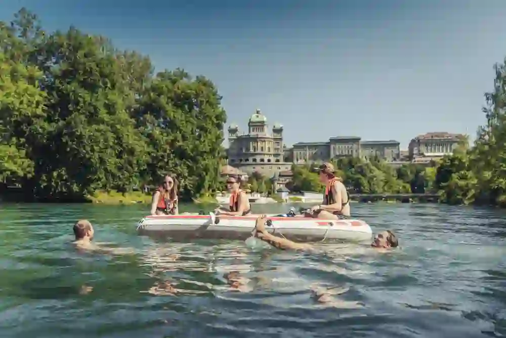 Un groupe de personnes joyeuses et un chien nagent et dérivent sur un bateau pneumatique sur une rivière, avec en toile de fond un décor urbain impressionnant, ce qui donne un sentiment de liberté et de plaisir estival.