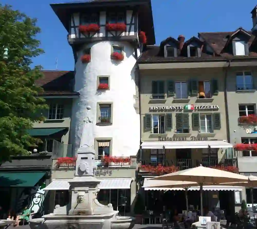 L'immagine mostra un edificio tradizionale europeo con un ristorante al piano terra, decorato con gerani rossi alle finestre. Di fronte al ristorante si trova una terrazza all'aperto con ombrelloni, che invita a soffermarsi.