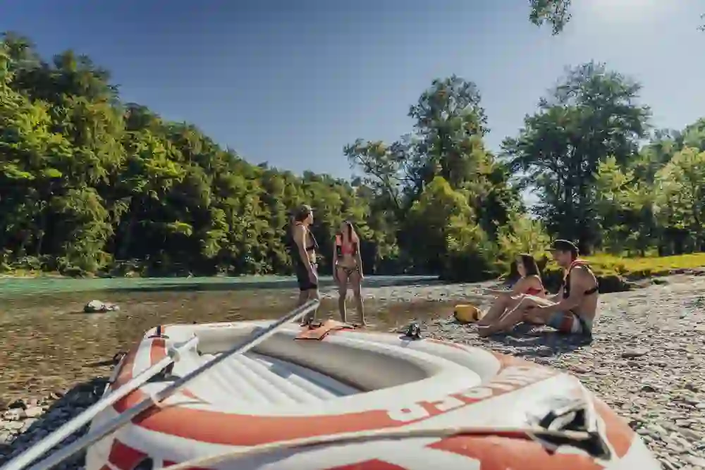 Eine entspannte Gruppe sitzt und steht am Kiesstrand der Aare nach einer Bootsfahrt. Sie geniessen die Sonne und die frische Luft, während das leere Schlauchboot neben ihnen liegt, Zeuge ihres jüngsten Abenteuers auf dem Wasser. Im Hintergrund zeichnet der Fluss eine natürliche Grenze durch das satte Grün des Uferwaldes.
