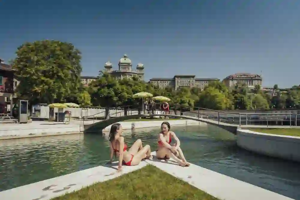 Due amici siedono sul bordo dell'acqua della piscina Marzili, ridendo e condividendo una conversazione rilassata, mentre il magnifico Palazzo Federale troneggia sopra di loro. Un'immagine della spensieratezza e dell'unione estiva a Berna.
