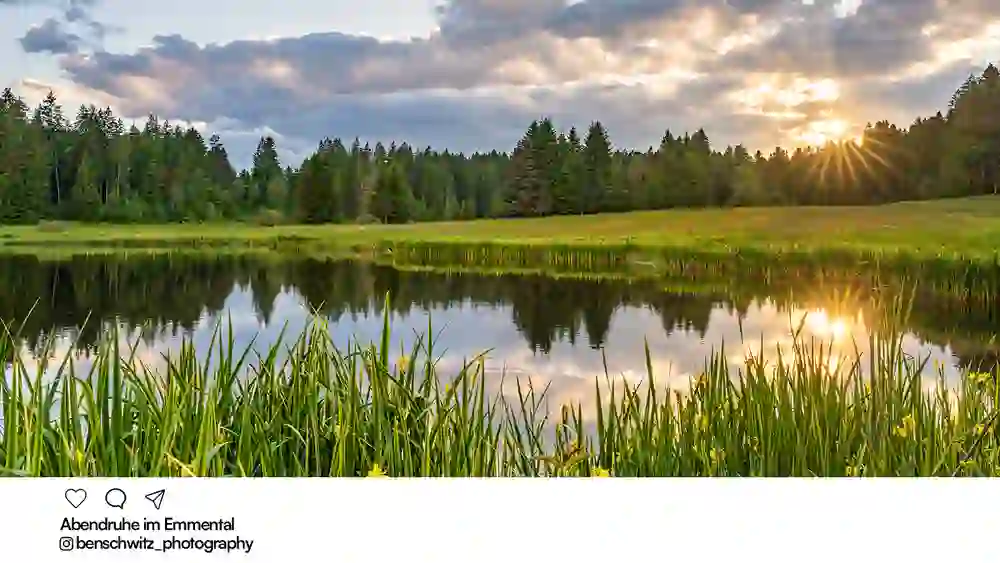 Un étang tranquille dans l'Emmental, entouré de prairies et de forêts. Le soleil couchant perce les nuages et se reflète dans l'eau - ambiance paisible et chaleureuse le soir.