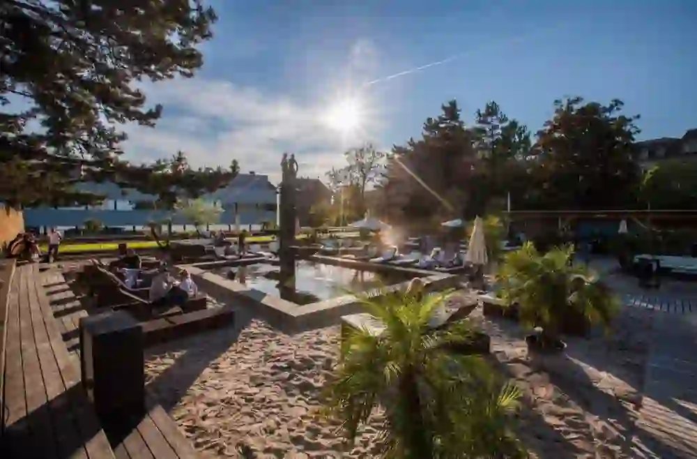The picture shows an urban oasis in Bern - the pop-up bar Summer Beach on the Grosse Schanze, which seems to bring the beach to the city. Rays of sunlight flood the scene and reflect on the water, while palm trees and sand give the place a tropical flair. Visitors relax on deckchairs and in lounge areas, enjoying the sun and the carefree atmosphere.