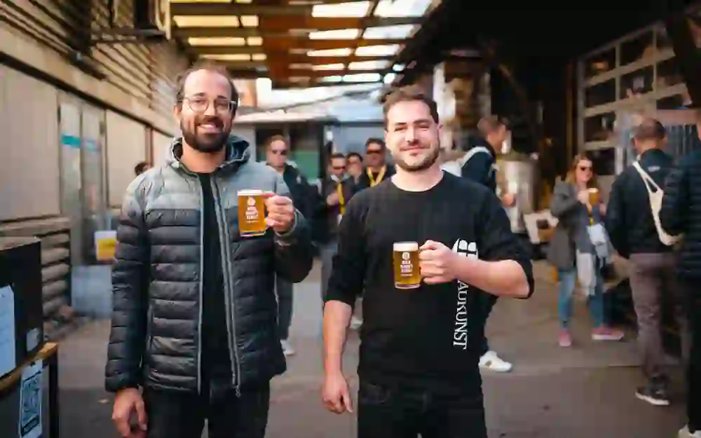 Das Bild zeigt zwei Männer, die Bierkrüge hochhalten und lächeln. Der linke Mann trägt eine Brille und eine schwarze Jacke, der rechte Mann ein schwarzes T-Shirt mit weißer Aufschrift. Im Hintergrund sind weitere Personen zu sehen, die ebenfalls Bier trinken. Die Szene spielt sich in einem überdachten Außenbereich a