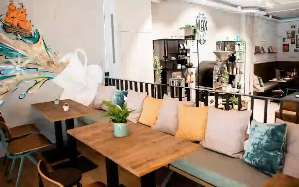 The picture shows the inviting interior of the Max coffee house, with cosy seating furnished with colourful cushions. Wooden tables add a rustic touch. A creative mural on the wall catches the eye, which together with the modern coffee bar creates a dynamic and stylish atmosphere. The ambience is relaxing and inviting, ideal for coffee lovers and those who want to enjoy a social moment.