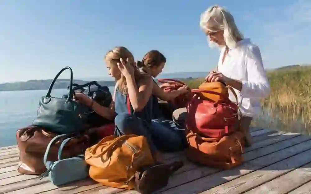 Tre generazioni di donne, riunite sulle rive di un lago, condividono un momento di tranquillità. Sono circondate da borse in pelle di alta qualità che riflettono il loro mestiere e il loro legame.