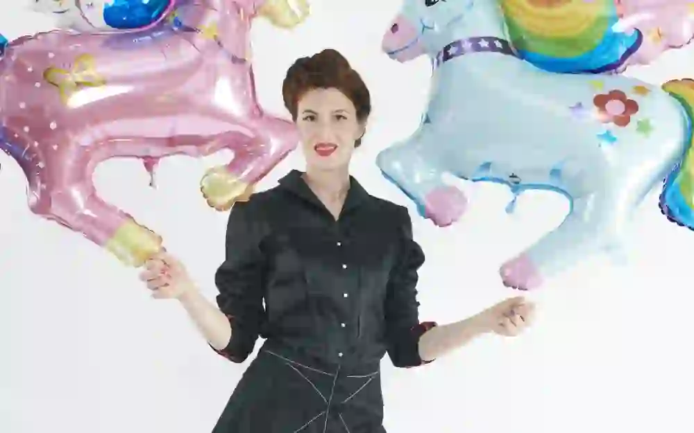 The woman in the picture stands smiling, surrounded by colourful helium balloons in the shape of stars and unicorns, creating a cheerful and festive atmosphere. Her pose and expression convey a mixture of professionalism and playfulness.