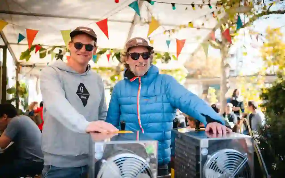 Das Bild zeigt zwei Männer in einem festlich dekorierten Bereich. Beide tragen Sonnenbrillen und Mützen. Der Mann links trägt einen grauen Pullover, der rechts einen blauen. Sie stehen vor zwei Lüftungsgeräten. Im Hintergrund sind bunte Wimpel und Menschen zu sehen.