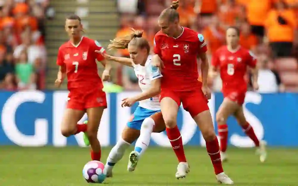 L'image montre un moment d'un match de football féminin où une joueuse en maillot blanc se bat pour le ballon contre deux adversaires en maillot rouge.