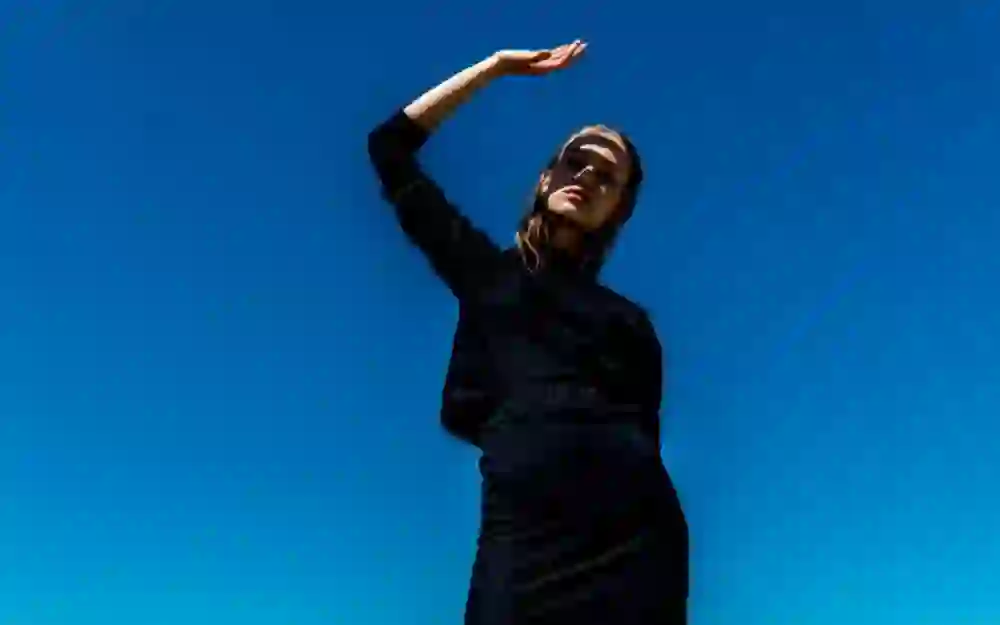 L'immagine mostra una donna in abito nero che tiene la mano davanti al viso per proteggerlo dal forte sole. Il suo sguardo è pensieroso, quasi sensuale, sotto il cielo azzurro e limpido.