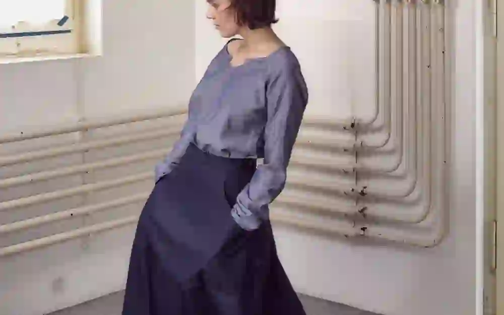 The photo shows the designer Rebora Rentsch in an elegant outfit, consisting of a grey top and a dark skirt, in front of an industrial backdrop with visible heating pipes. The pose and the contemplative look convey a calm and thoughtful mood.