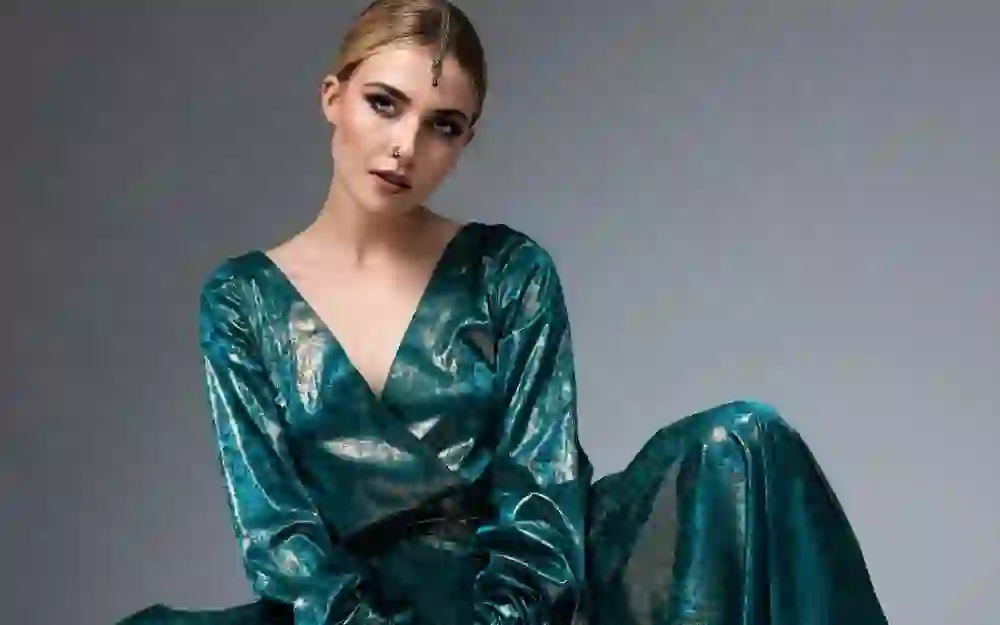 The picture shows a woman in a shiny turquoise dress that radiates elegance and sophistication. Her posture is confident and her intense gaze gives the picture a strong presence. The plain grey background emphasises her striking appearance.