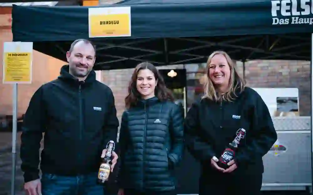 Das Bild zeigt drei Personen vor einem schwarzen Zelt mit der Aufschrift "Felsenau". Der Mann links hält eine Bierflasche und trägt eine schwarze Jacke. In der Mitte steht eine lächelnde Frau mit braunen Haaren, rechts eine weitere Frau mit blonden Haaren, die ebenfalls eine Bierflasche hält.