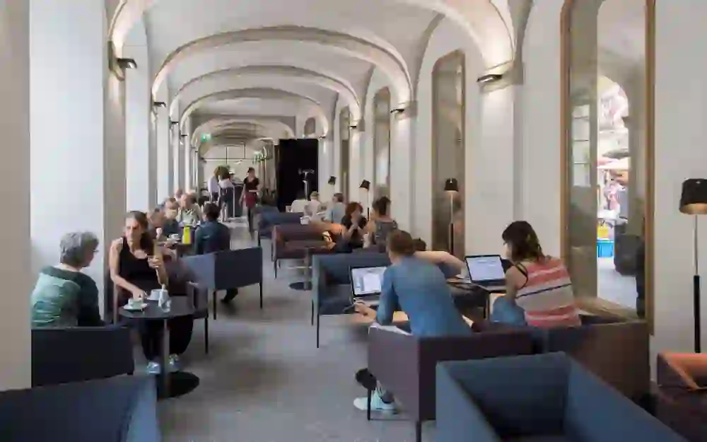 Das Bild zeigt das lebendige Treiben in der Café-Lounge Lesbar in Bern. Unter den gewölbten Arkaden sitzen Menschen in tiefen Gesprächen, während andere in die Welt ihrer Laptops versunken sind. Die moderne Einrichtung unter historischer Architektur schafft eine Atmosphäre, die sowohl zur Kollaboration als auch zum individuellen Schaffen einlädt.