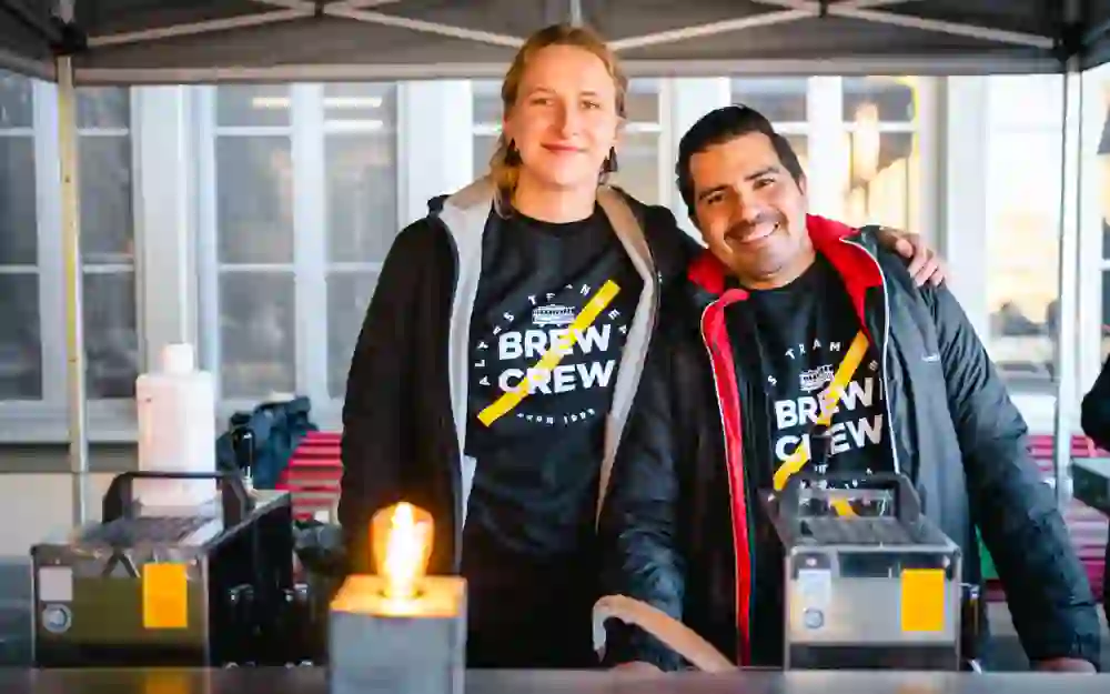 Das Bild zeigt zwei Personen, eine Frau und einen Mann, die hinter einem Tisch stehen und lächeln. Beide tragen schwarze T-Shirts mit der Aufschrift "Brew Crew" und sind in dunklen Jacken gekleidet. Vor ihnen stehen zwei Maschinen und eine brennende Kerze. Die Szene spielt sich in einem überdachten Bereich ab, im Hintergrund sind Fenster zu sehen.