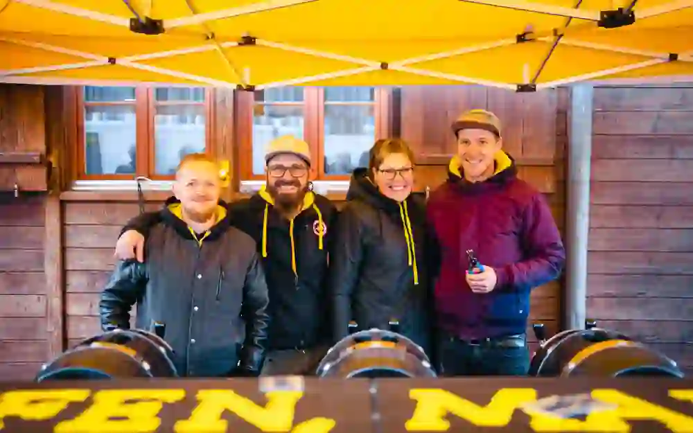 Das Bild zeigt vier Personen, die unter einem gelben Zelt vor einer Holzwand stehen. Alle tragen Jacken, die drei Männer auch Mützen. Sie lächeln und haben die Arme umeinander gelegt. Vor ihnen stehen zwei Bierfässer mit Zapfhähnen. Das Zelt hat die Aufschrift "OPEN NOW".