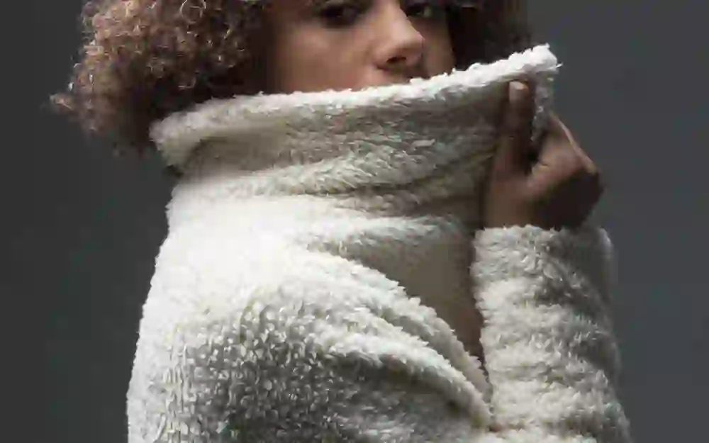 L'immagine ritrae la stilista Etris in un soffice capo bianco che emana intimità e calore. Lo sguardo gentile e la postura che sfiora delicatamente il materiale suggeriscono un momento di calma e fiducia in se stessi.