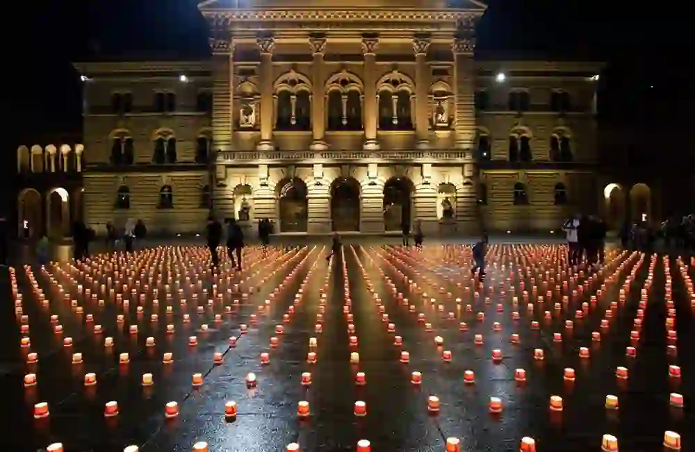 Das Bild zeigt den Bundesplatz, erleuchtet von hunderten Kerzen, die eine ruhige und feierliche Atmosphäre schaffen. Menschen gehen staunend zwischen den Kerzen entlang, während das beleuchtete Bundeshaus im Hintergrund majestätisch erscheint.