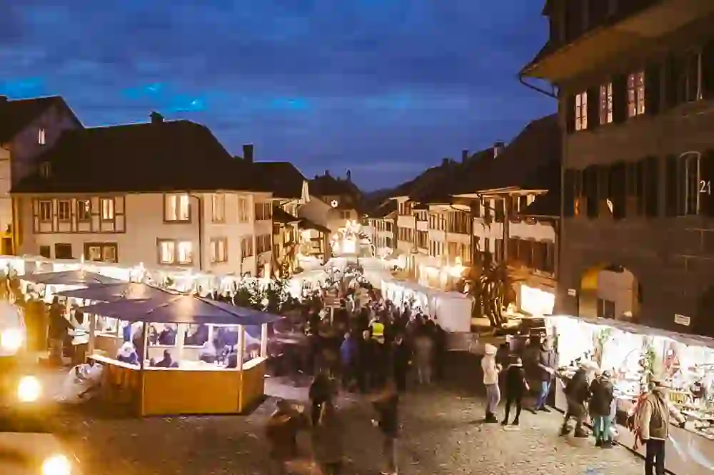 Das Bild zeigt einen belebten Weihnachtsmarkt in einer malerischen Altstadt bei Einbruch der Dämmerung. Die festlich geschmückten Stände und die erleuchteten Häuserfronten strahlen eine warme und einladende Atmosphäre aus. Die Besucher genießen das gemütliche Treiben und die festliche Stimmung.