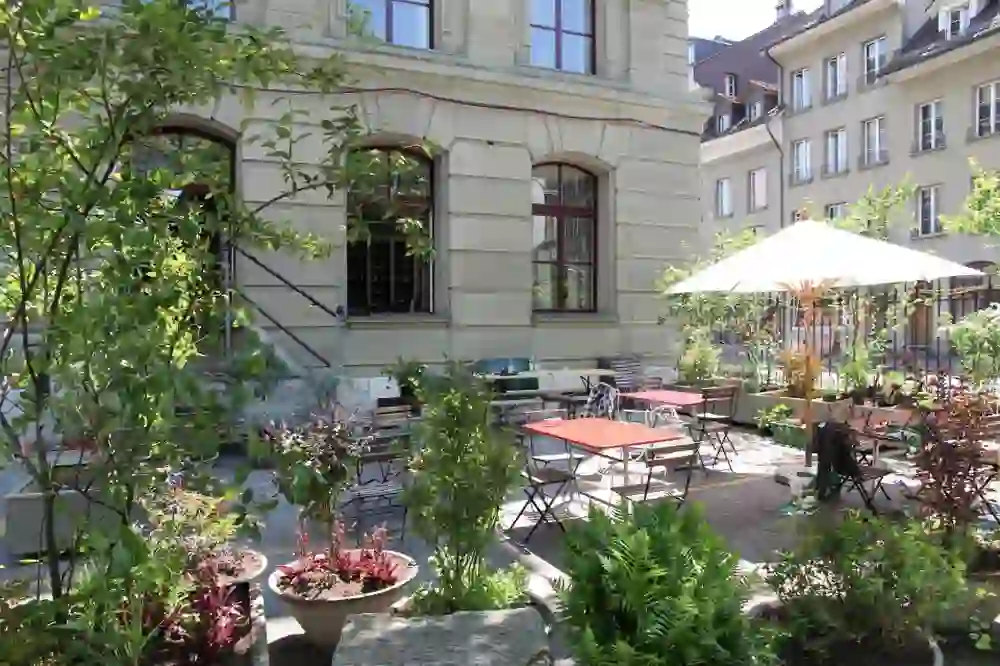 The picture shows a cosy garden terrace of the staff room in Bern. The weather is sunny. You can see a white parasol providing shade. The terrace is decorated with many green plants
