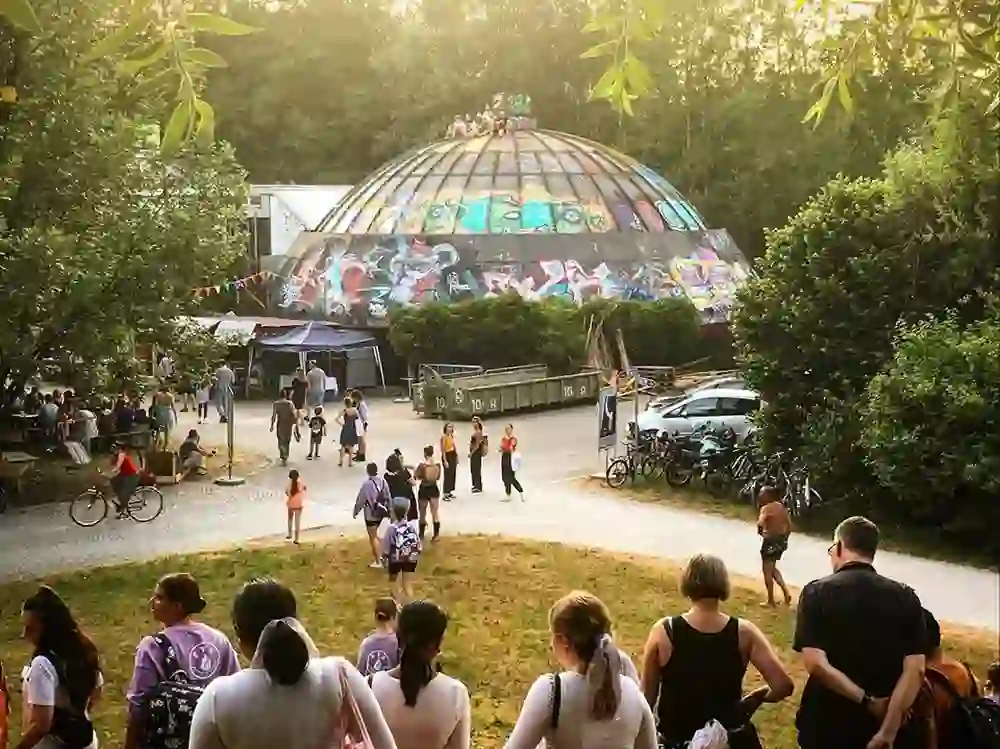 L'image montre le Gaskessel à Berne, vivement décoré de graffitis, avec en arrière-plan un lieu de rencontre animé. Des personnes d'âges différents se promènent ou s'attardent, ce qui suggère une atmosphère conviviale et détendue. Cela donne l'impression d'un melting-pot culturel dans la chaude lumière du soir.