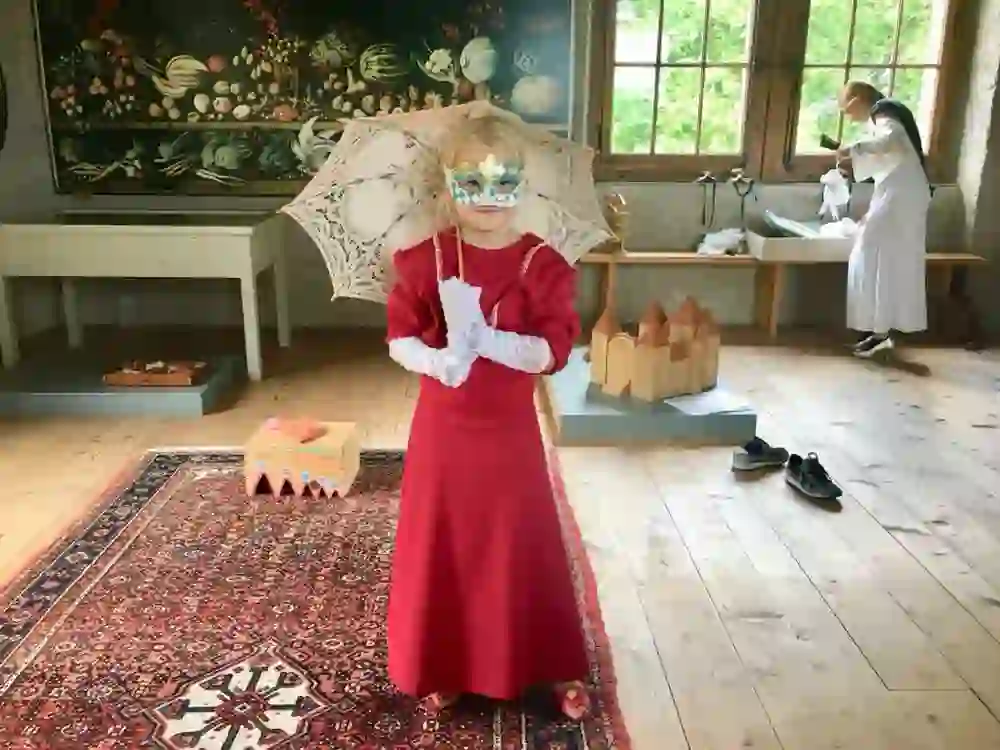 Ein Kind, verkleidet in historischer roter Robe und weisser Maske, posiert in einem Zimmer mit historischer Atmosphäre, untermalt von spielerischer Neugier und dem Charme vergangener Zeiten.