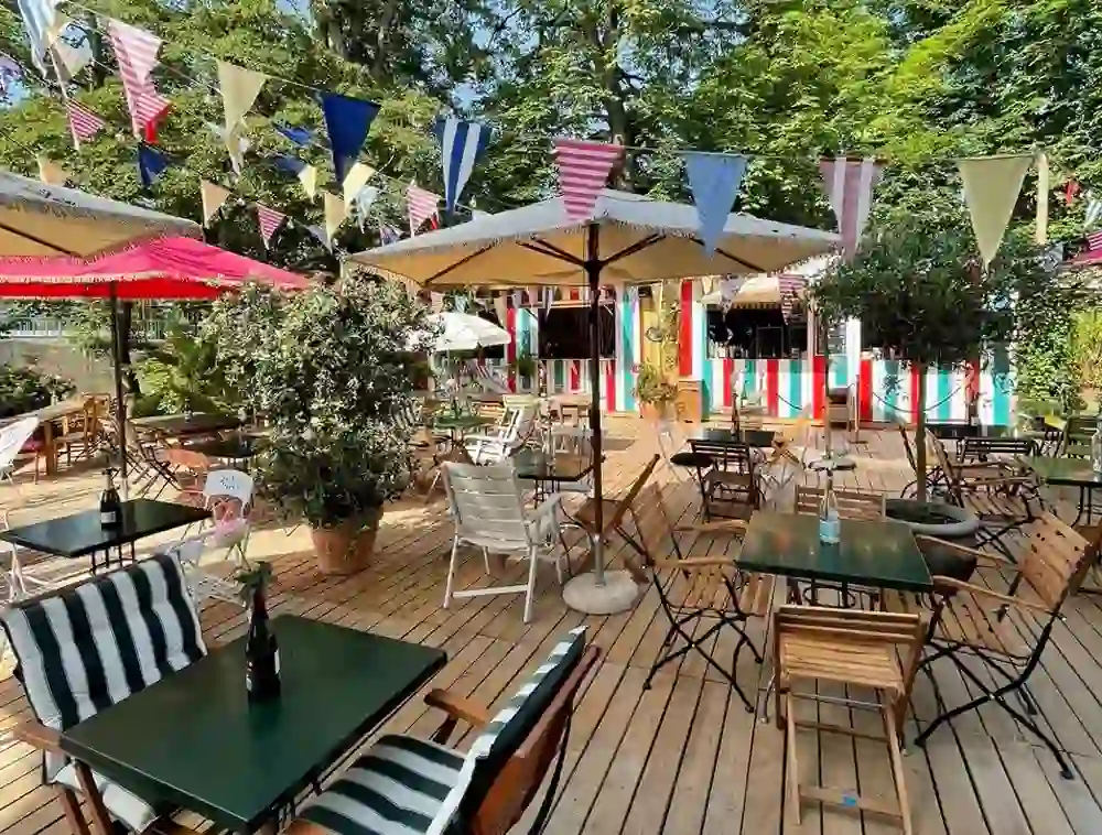 Das Bild strahlt eine friedliche und einladende Atmosphäre aus und zeigt eine Pop-up-Bar im Freien, „Park am Wasser“ in Bern. Die sommerliche Terrasse ist mit bunten Wimpelketten geschmückt und lädt zum Verweilen ein. Unterschiedliche Sitzgelegenheiten bieten für jeden Geschmack einen Platz an der Sonne oder im Schatten großer Schirme und Bäume. Die warmen Holzfarben der Tische und Stühle auf dem Holzdeck verleihen dem Ort eine natürliche und entspannte Atmosphäre, perfekt für ein Getränk in der Sommersonne oder für gesellige Stunden mit Freunden.