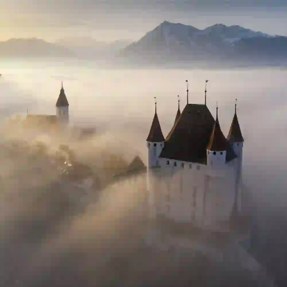Das Bild zeigt eine mittelalterliche Burg, die aus dem Nebel ragt. Die Burg hat mehrere Türme mit spitzen Dächern. Im Hintergrund sind Berge zu sehen, die ebenfalls teilweise im Nebel verborgen sind. Die Atmosphäre wirkt mystisch und ruhig.