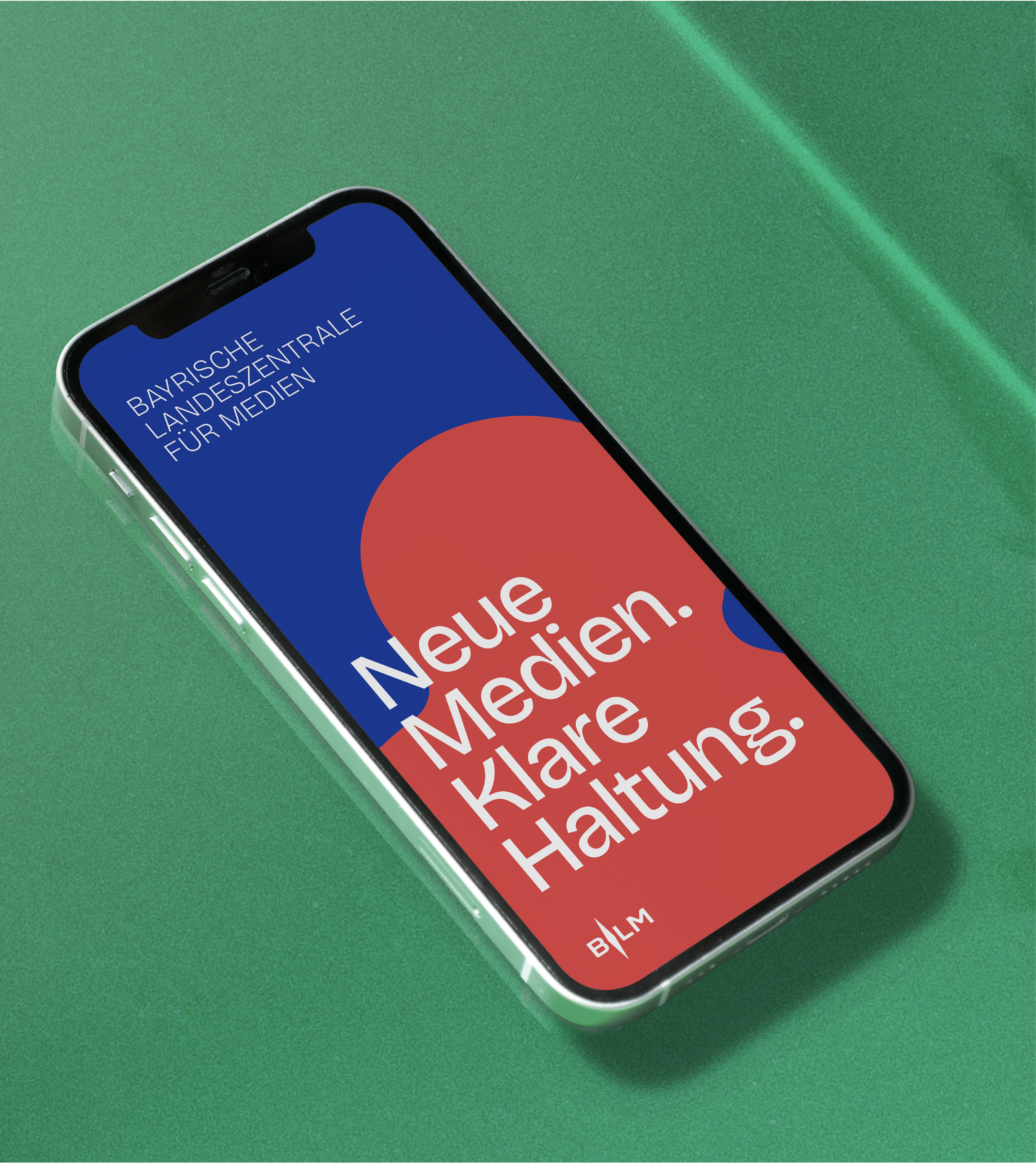 Bayerische Landeszentrale für neue Medien Ein Handy auf einem grünen Untergrund. Am Screen ist ein modernes Plakatdesign der Bayerischen Landeszentrale für Medien mit klarer Typografie und kontrastreichen Farben in Blau und Rot.
